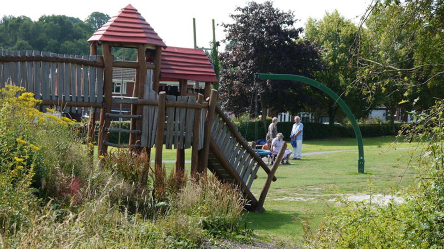 Wooden treehouse climbing frame in foreground, people sitting on bench in distance