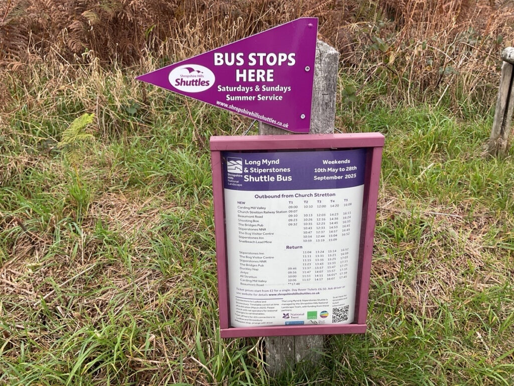 Photo showing the bus stop signage at The Knolls which has the Shropshire Hills National Landscape's Shuttle Bus Flag and Time Table.