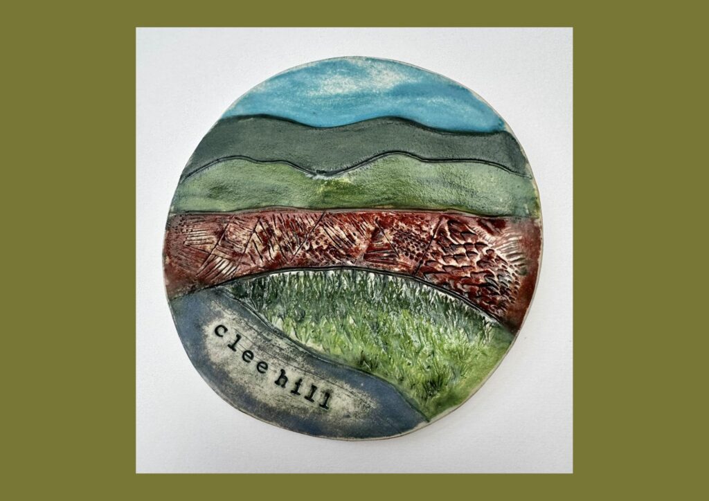 An circular organic shaped ceramic tile is decorated with green brown and blue glaze with relief and textured designs that depict the hills, fields and sky. The words Clee Hill are embossed at the bottom of the tile.