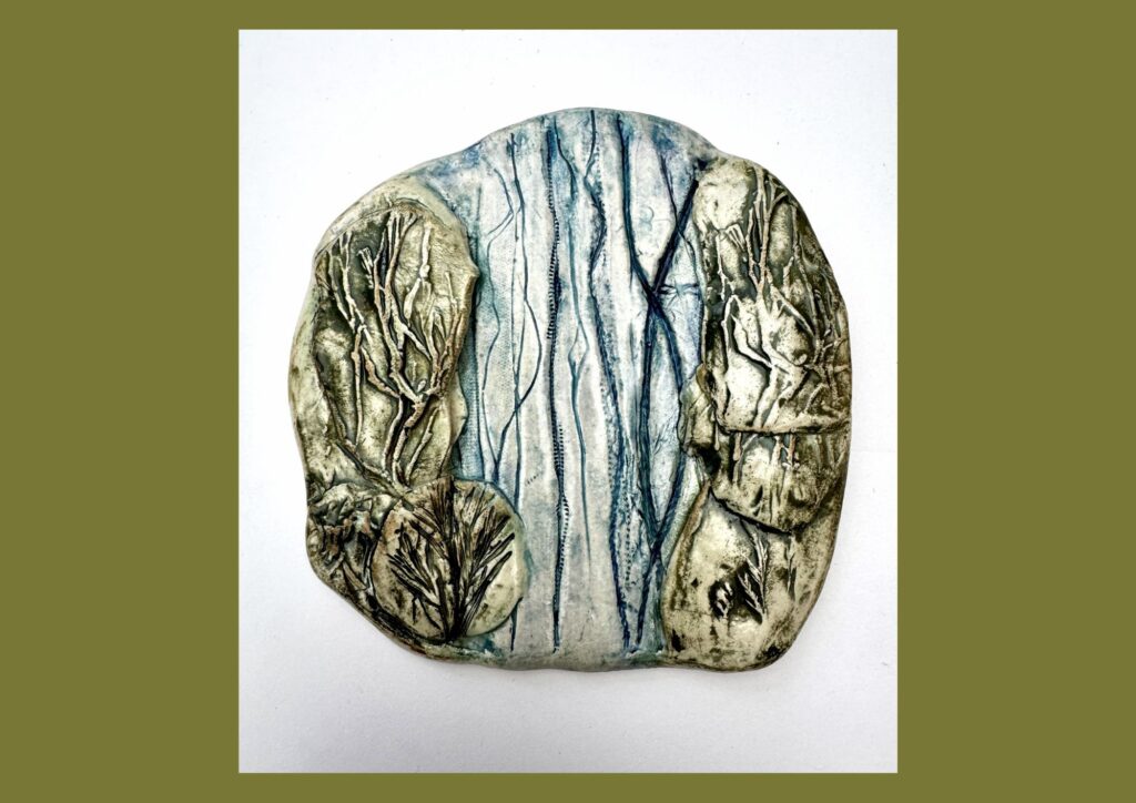 A round, organic shaped ceramic tile is decorated in muted green, brown and blue glazes and raised textured shapes to represent the landscape and brook.
