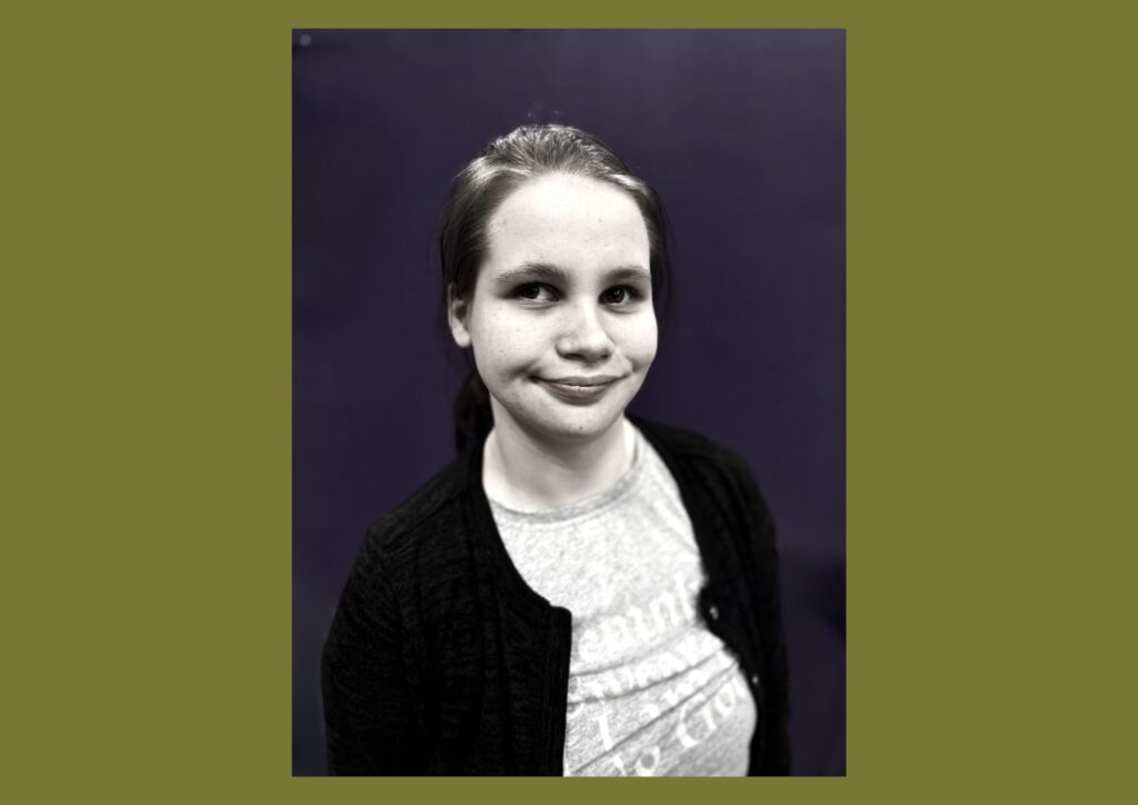 The artist is shown in a black and white image smiling at the camera. She is against a dark backdrop and wearing a black cardigan.