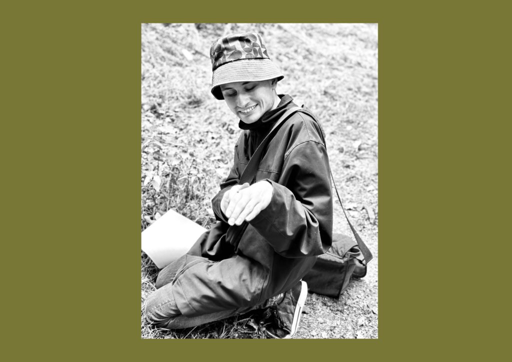 The artist is shown in a black and white image on outside and sat on the grass. They are wearing a hat and carrying a bag.
