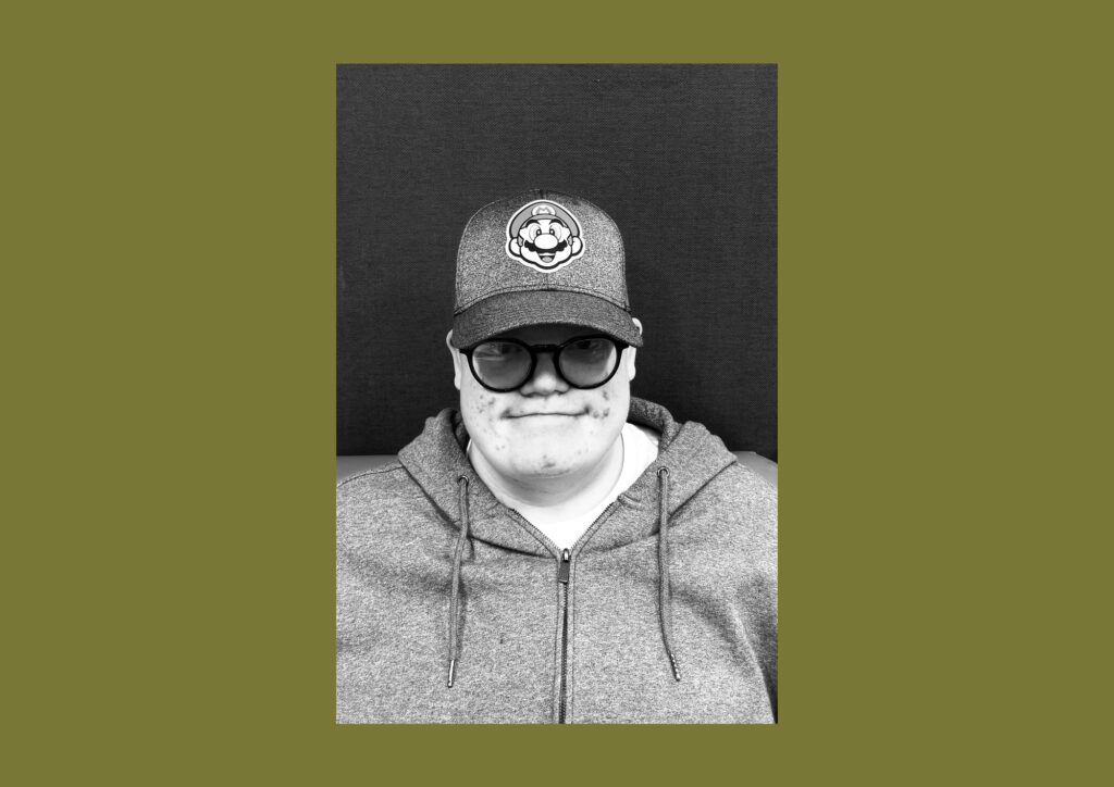 The artist is seen in a black and white photograph smiling at the camera. He is wearing glasses, a hoodie and a baseball cap. There is a black backdrop.