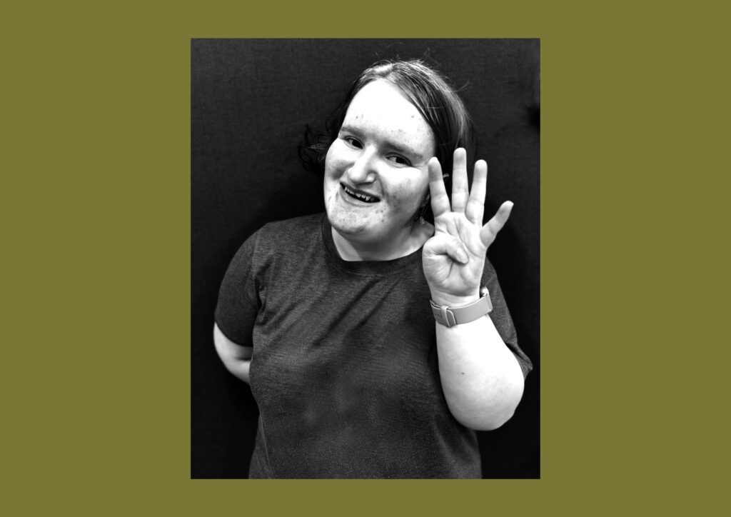 The artist is shown in a black and white image smiling at the camera wearing black clothes and holding four fingers up