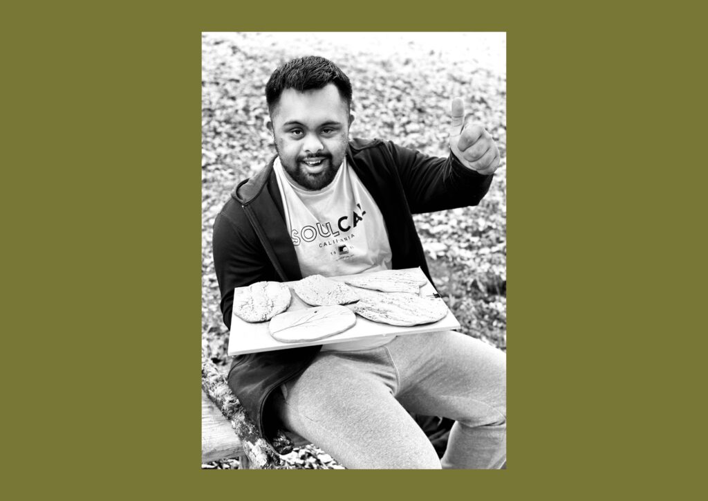 The artist is shown in a black and white image sat on the grass waiving at the camera and holding a tray of unfinished tiles.