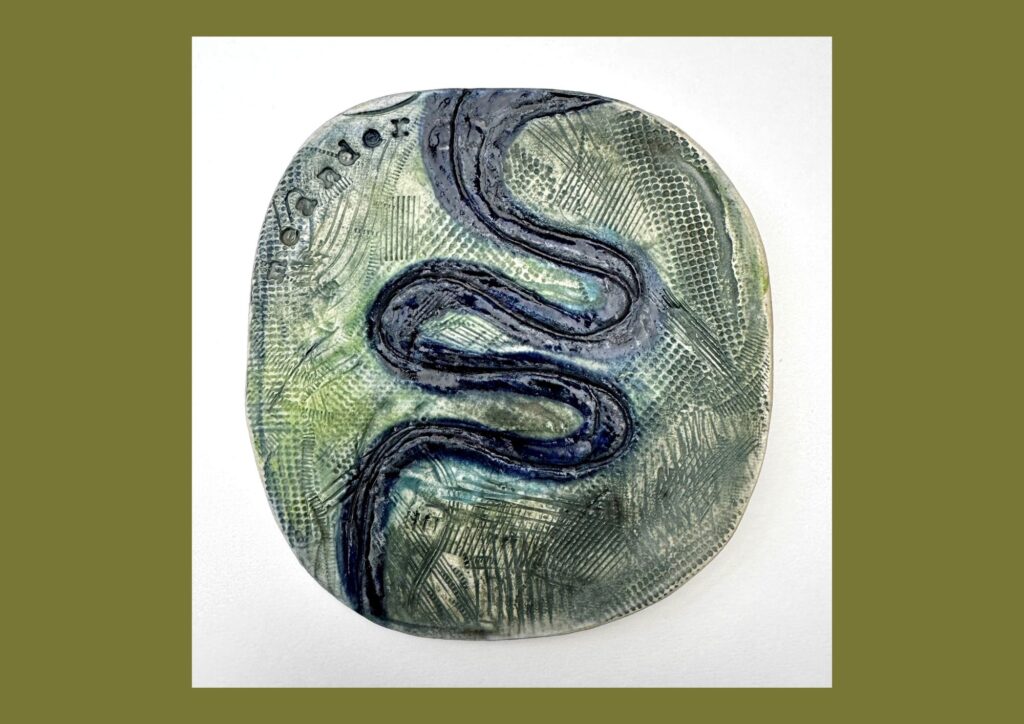 A circular organic shaped ceramic tile is decorated with green and blue glaze with the meandering line of blue stretching from the top to the bottom of the tile.