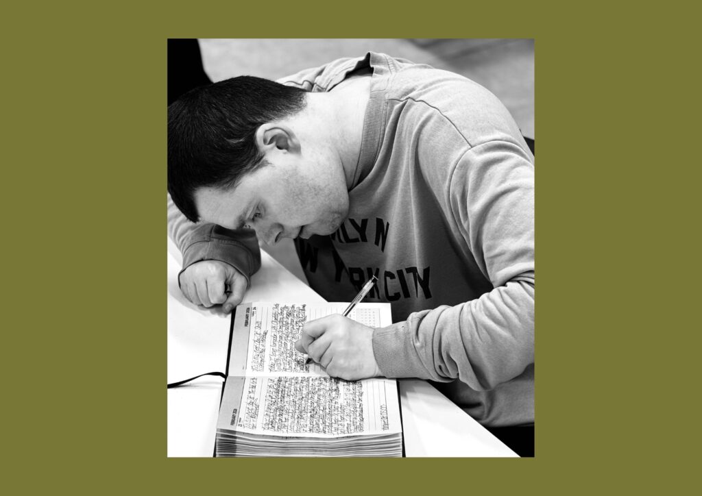 The artist is shown in a black and white image writing in a lined journal. He is looking directly at the book.