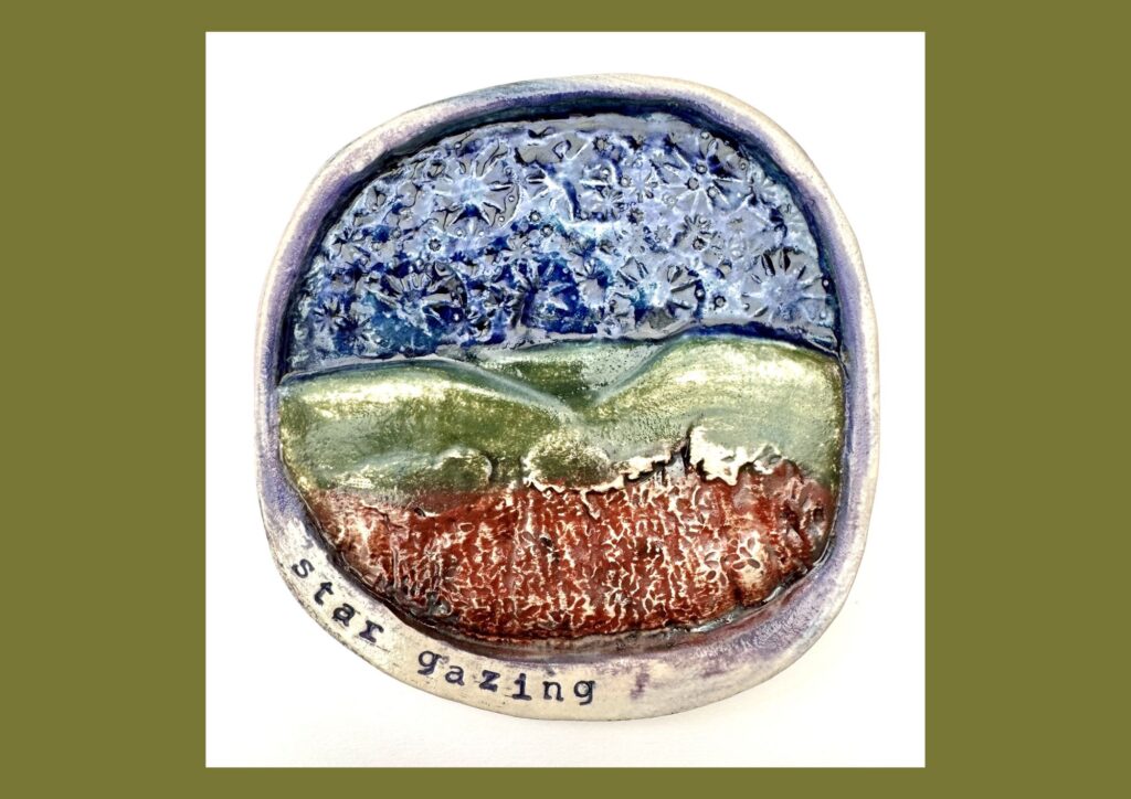 A circular organic shaped ceramic tile is decorated in brown, green and blue glaze with textured elements that depict the hills and sky. The words Star Gazing are embossed in the clay