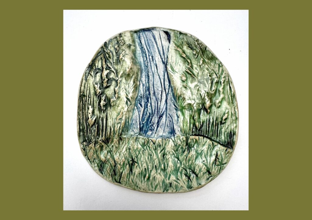 A circular organic shaped ceramic tile, is decorated with green, blue and brown glazes and textured patterns to represent the hills and water in the landscape.