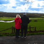 A woman in a red coat and a man in a black coat look out over a valley and towards a flood plain where the river meanders.