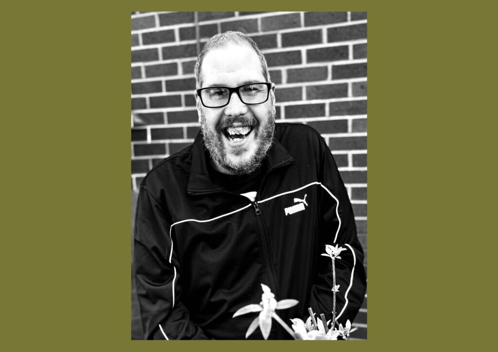 The artist is shown in a black and white photograph, smiling at the camera. He is outside with a brick wall backdrop.