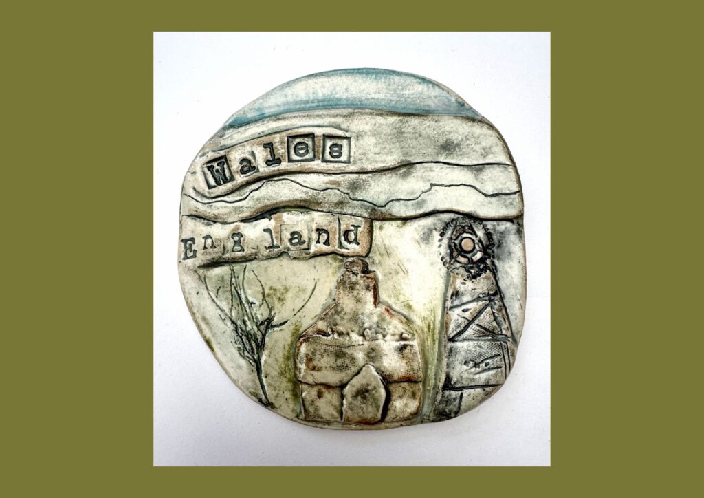 A round organic shaped tile is decorated with green, blue and brown glaze, with raised and textured designs that show the hills, sky and old pit head gear.