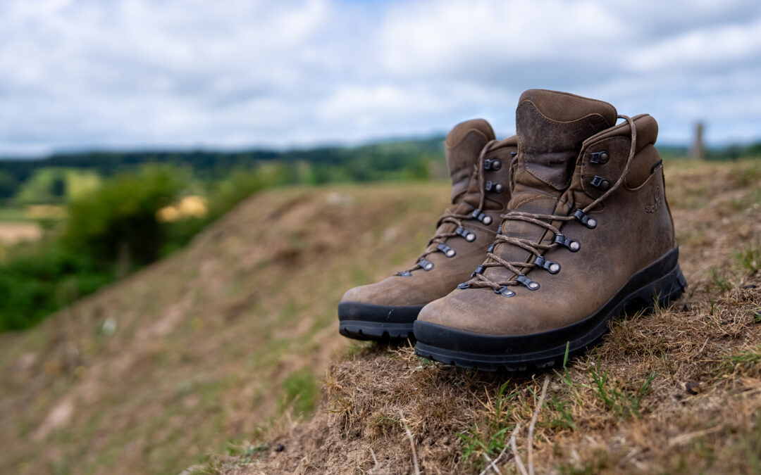 A pair of walking boots is left on the side of a grass bank. In the distance are hills and trees. The setting would be a wonderful place for a walk