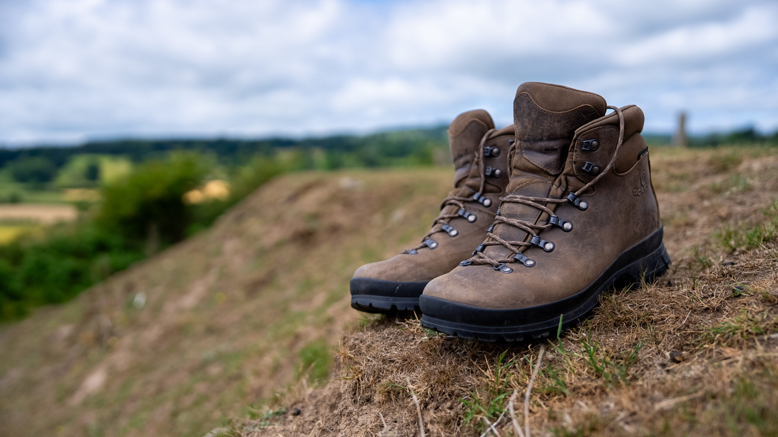 A pair of walking boots is left on the side of a grass bank. In the distance are hills and trees. The setting would be a wonderful place for a walk